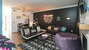 A modern living room with a black and white geometric rug, a black leather sofa with colorful cushions, a black coffee table with books and a lamp, and a neon sign on the wall. - 90's in Nash! 2bd Townhome in WEST END! 8 min ->DT! (Nashville)