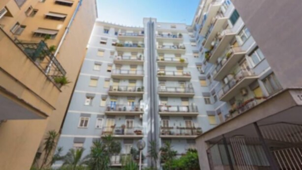 A high-rise residential building with multiple balconies, a central elevator shaft, and surrounding greenery.
