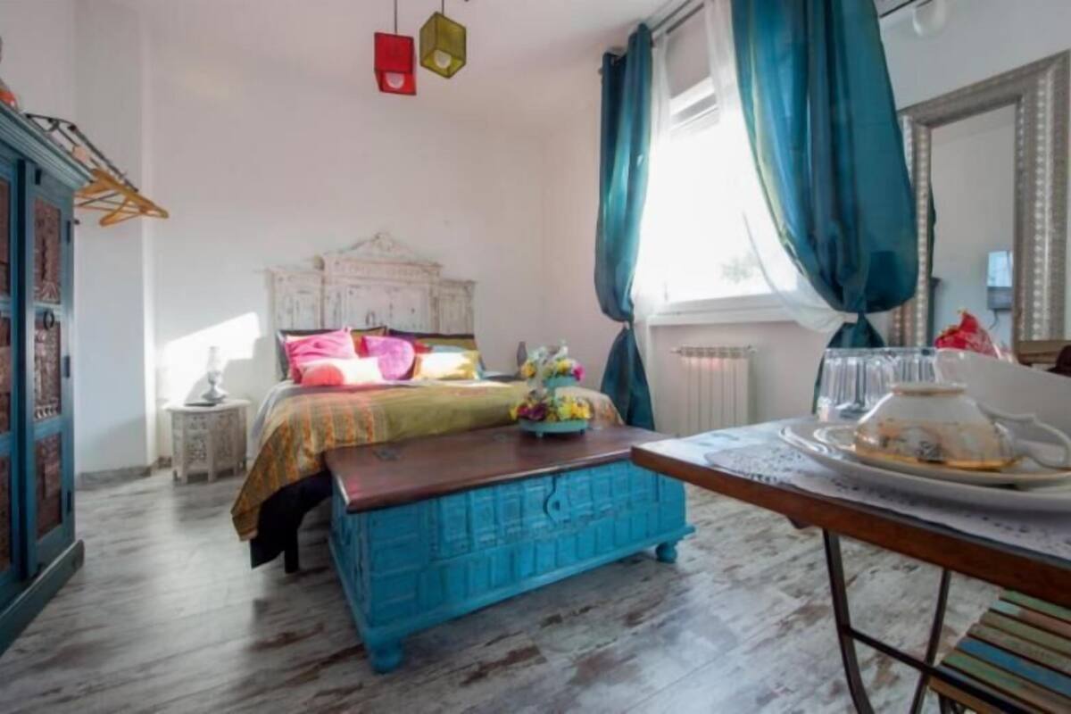 A bedroom with a bed, a dresser, a table with a teapot, and a window with curtains.