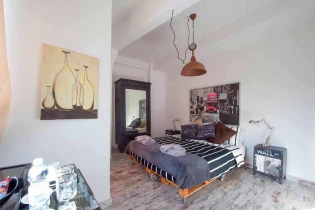 A bedroom with a bed, a mirror, a wall art piece, a lamp, and a small table with water bottles.