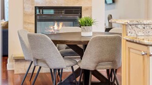 Un espace de salle à manger moderne avec une table centrale, des chaises à revêtement gris, un lustre contemporain, une cheminée et une œuvre d’art encadrée au mur.
