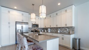 A modern kitchen with white cabinets, a stainless steel refrigerator, a microwave, and a large island with bar stools. - NEW 3BR Luxury Townhome (Heber City)