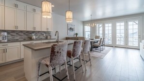 A modern kitchen with white cabinets, a granite countertop, and a central island with bar stools. There are pendant lights above the island and a dining area with a view of the outside. - NEW 3BR Luxury Townhome (Heber City)