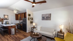 Una cocina moderna con armarios de madera oscura, un refrigerador negro y una encimera blanca. Hay una zona de comedor con un sofá blanco, un sillón amarillo y una mesa de centro redonda.