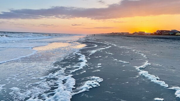 Une plage avec des vagues, un coucher de soleil et des bâtiments en arrière-plan.
