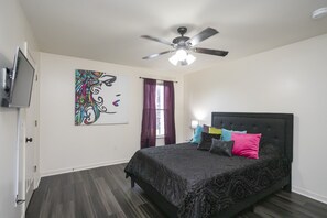 A bedroom with a bed, a ceiling fan, a TV, a window with curtains, and a wall art piece.