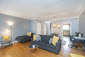 A living room with a grey sofa, a small coffee table, and a bench. There is a desk with a chair and a plant. The room has wooden flooring and light blue walls.