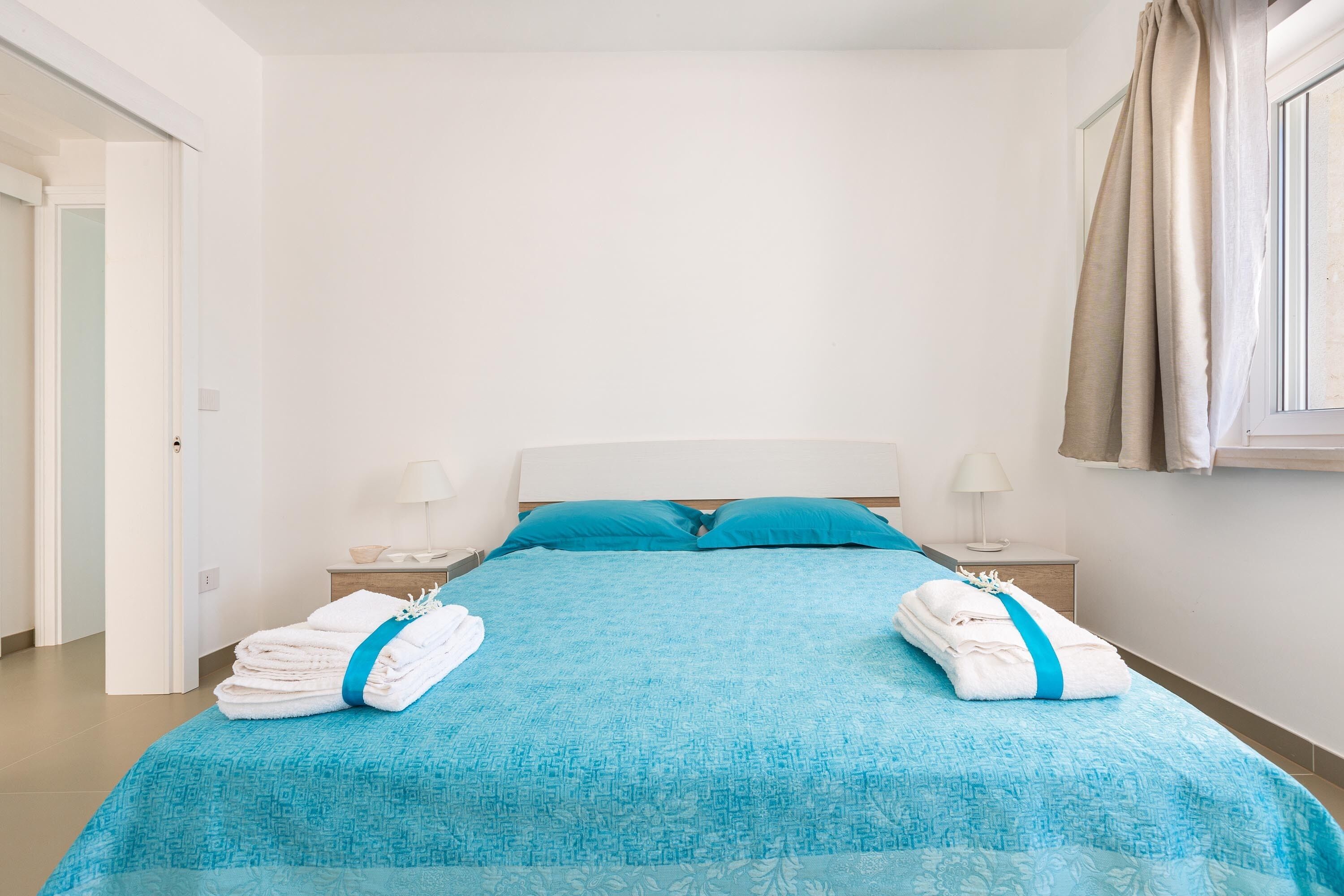 A neatly made bed with a blue bedspread and pillows, two folded towels with blue ribbons, a bedside table with a lamp, and a window with curtains.