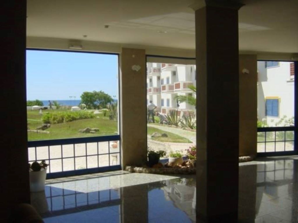 Eine großzügige Lobby mit einer großen Glastür, die auf einen Balkon mit Blick auf einen angelegten Garten und Gebäude in der Ferne führt.