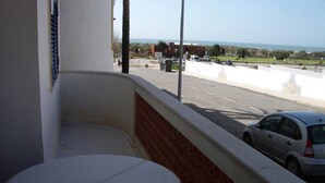 A balcony with a round table, a view of a road with cars, and a building in the background. - Robin Apartment, Manta Rota, Algarve (Vila Nova de Cancela)