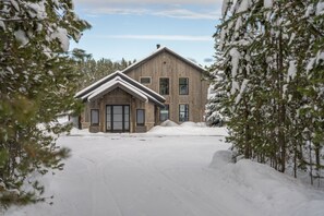 A wooden cabin with a gabled roof, surrounded by snow-covered ground and trees.