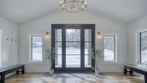A spacious entryway with a wooden floor, a bench, a chandelier, and a view of a snowy landscape through large windows. - Shoreline Lodge, Lakefront and hot tub (Island Park)