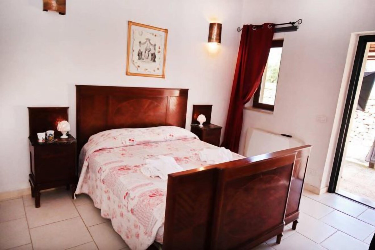 A bedroom with a wooden bed, a nightstand, a bedside table, a wall-mounted lamp, a window with red curtains, and a framed picture on the wall.