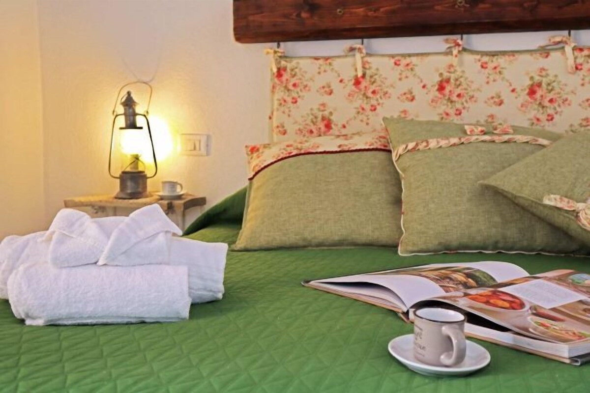 A neatly made bed with green bedding, a floral headboard, a bedside lamp, a stack of white towels, and a magazine with a cup of coffee on the side.