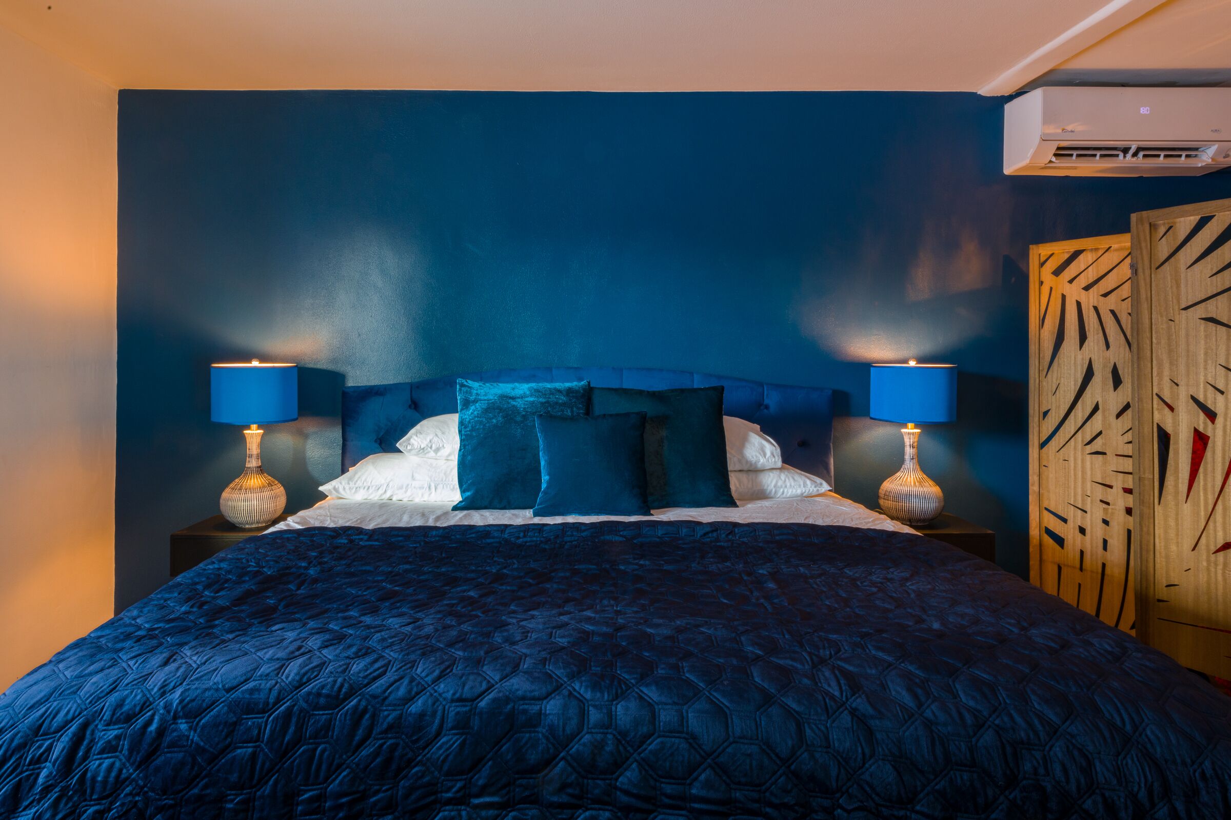 Camera da letto con parete blu scuro, un letto con testiera blu, due abat-jour e un pannello di legno con motivi geometrici.