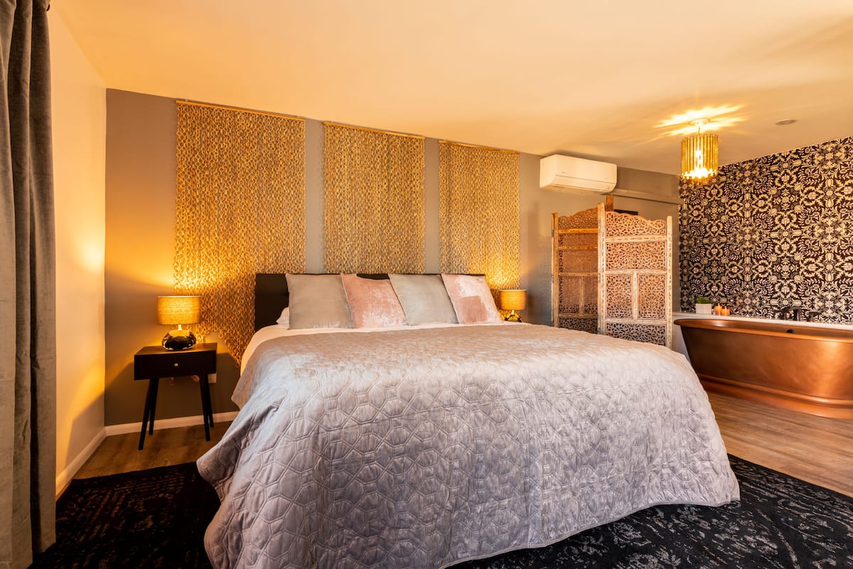 Ein Hotelzimmer mit Bett, Nachttisch, Lampe, Badewanne und Wandverkleidung mit Muster.