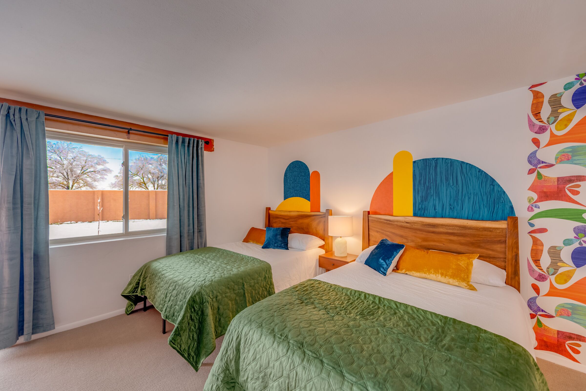 A hotel room with two beds, a window with curtains, a wall with colorful abstract art, and a wooden headboard.