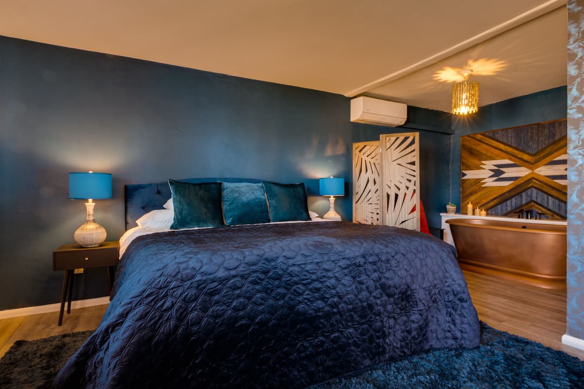 Ein modernes Schlafzimmer mit dunkelblauer Wand, einem großen Bett mit gesteppter Bettdecke, zwei Nachttischen mit Lampen, einem Holzkopfende und einer dekorativen Wandverkleidung sowie einer Kupferbadewanne.