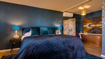 A modern bedroom with a dark blue wall, a large bed with a quilted duvet, two bedside tables with lamps, a wooden headboard, a decorative wall panel, and a copper bathtub.