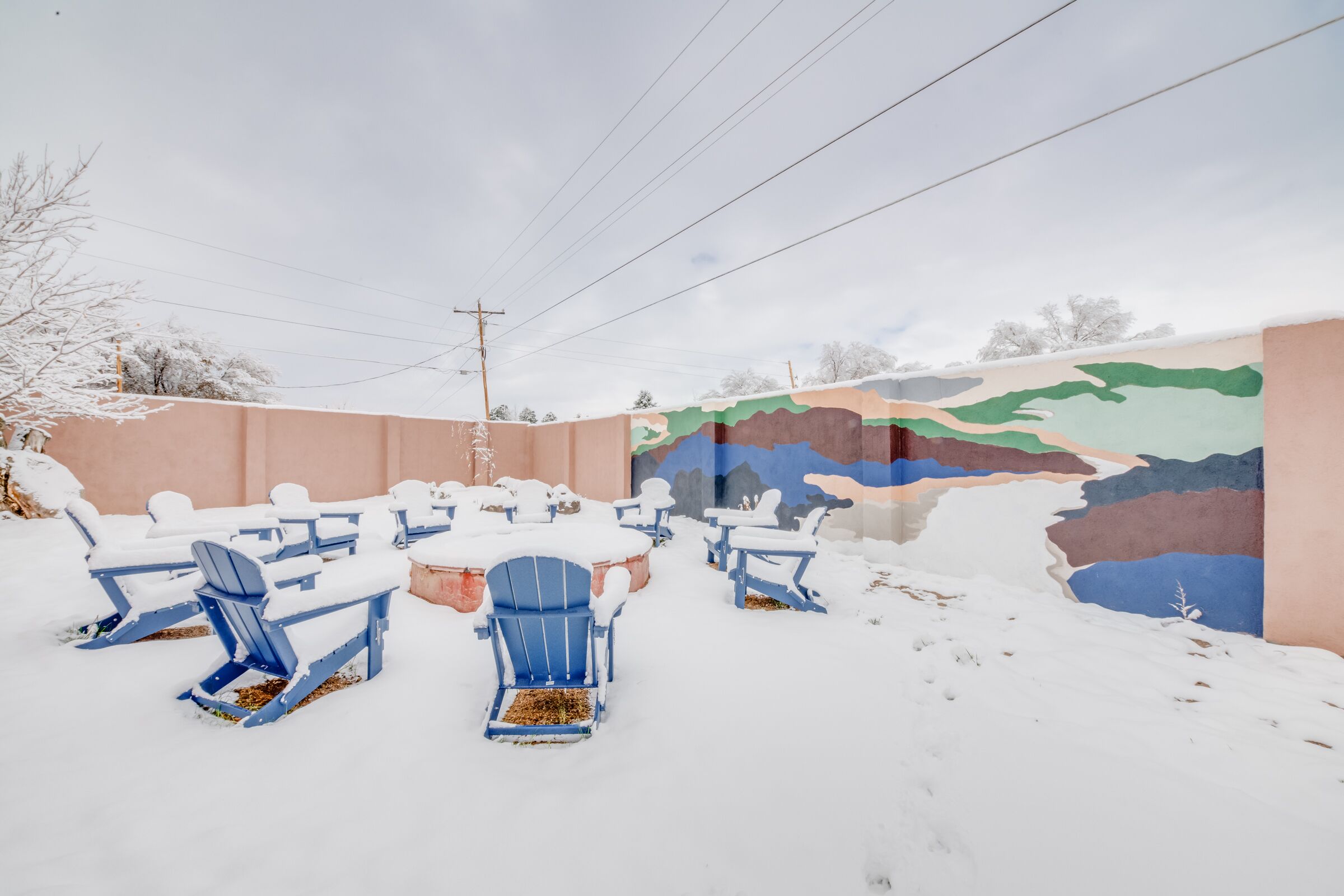 A snow-covered outdoor seating area with blue chairs and tables, a mural on the wall, and a power line in the background.