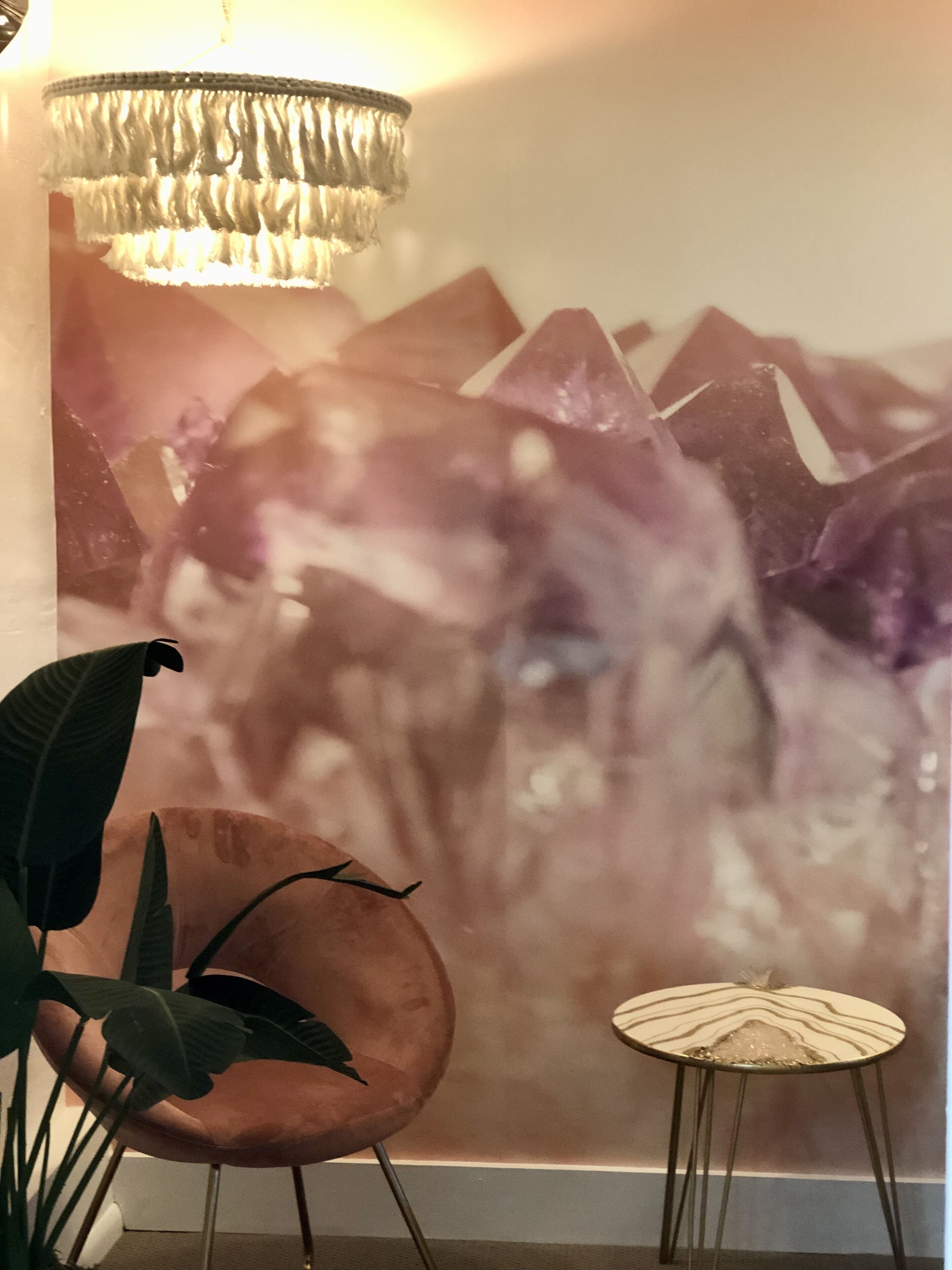 A room with a large abstract painting on the wall, a modern chair, a small round table, and a hanging light fixture.
