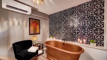 Une salle de bain moderne avec une baignoire sur pieds, un fauteuil en velours noir, une petite table avec des bougies et un mur à motifs.