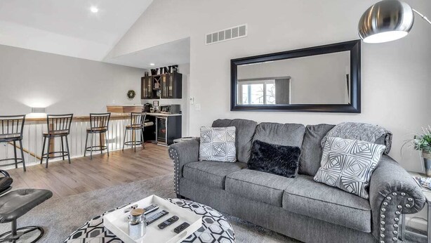 Una sala moderna con un sofá gris, una mesa de centro y una zona de comedor con taburetes de bar.