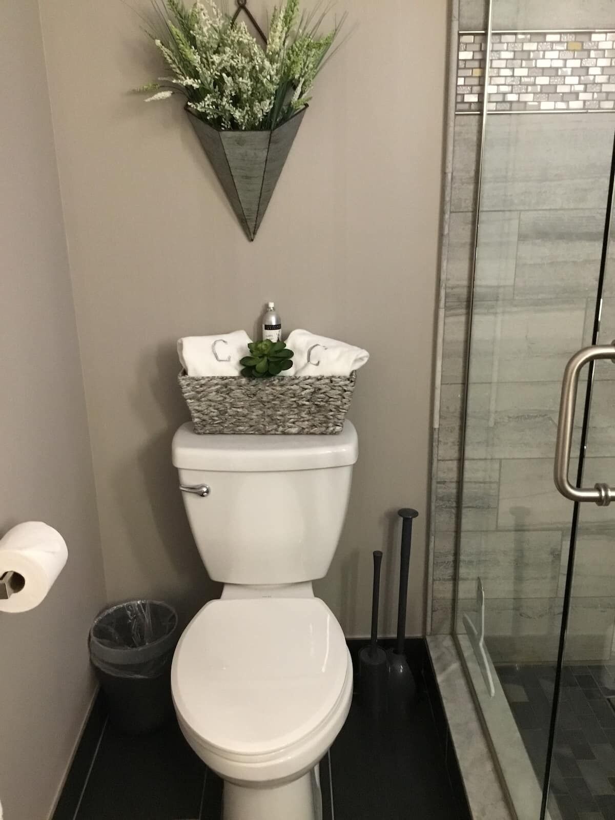 A bathroom with a toilet, a trash bin, a toilet paper roll, a shower area with a glass door, and a wall-mounted plant.