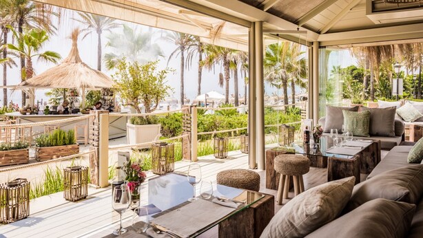 A sunlit outdoor seating area with a view of palm trees and a beach. - Magnificent recently built villa Pax 12 sea view Golden Mile Marbella (Marbella)