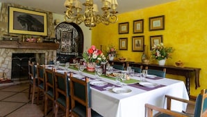 Un coin repas avec une longue table dressée pour le repas, des chaises, un lustre, une cheminée et des tableaux encadrés accrochés au mur.