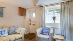 A cozy living room with a sofa, armchair, and a small table. A window with curtains and a view of a garden is visible. - 30 Sherborne Lane (Lyme Regis)