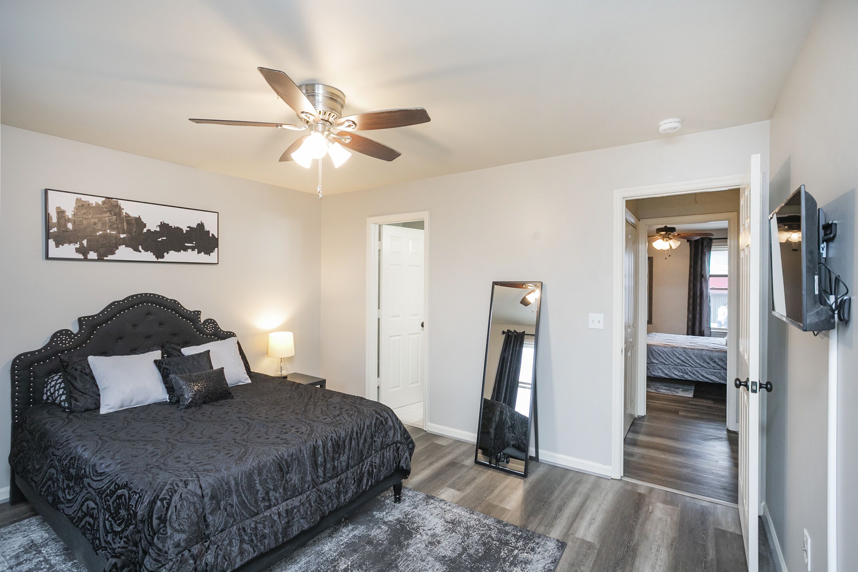 A bedroom with a bed, a ceiling fan, a mirror, and a door leading to another room.