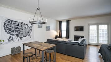 A modern living room with a wooden table, bar stools, a grey sofa, and a chandelier.