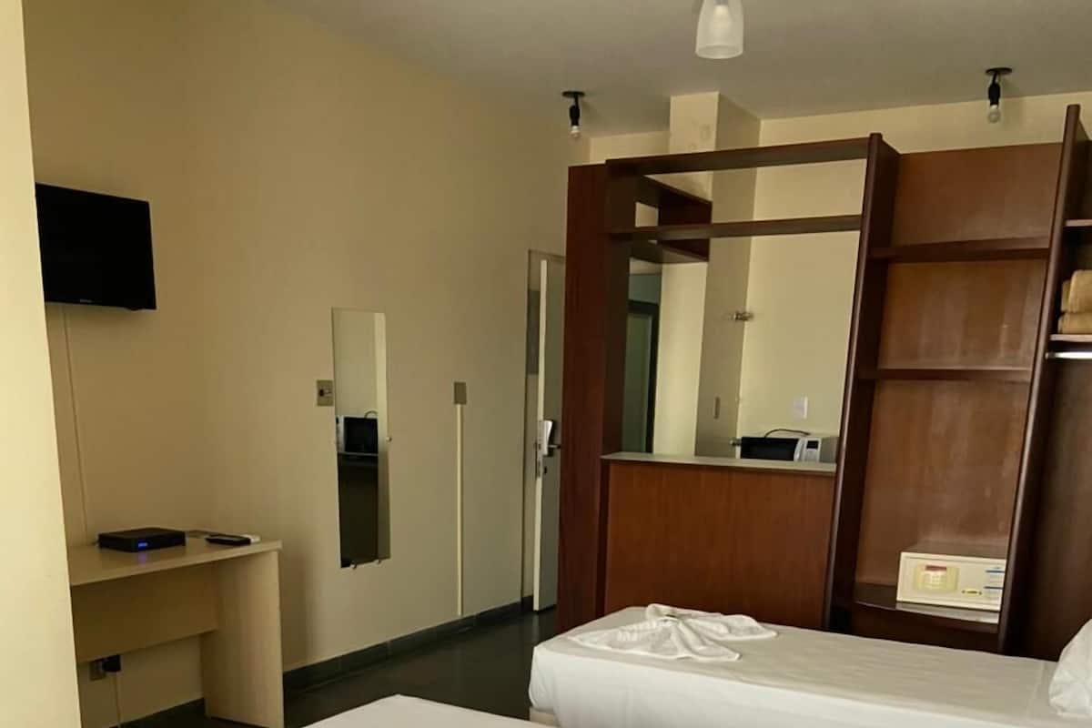 Ein Hotelzimmer mit zwei Betten, einem kleinen Schreibtisch, einem Sessel, einem Fernseher und einem Spiegel.