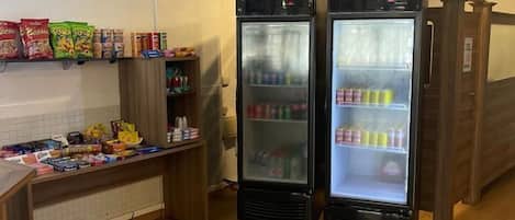 A room with two black refrigerators stocked with beverages, a wooden counter with snacks, and a wall-mounted shelf with packaged goods.