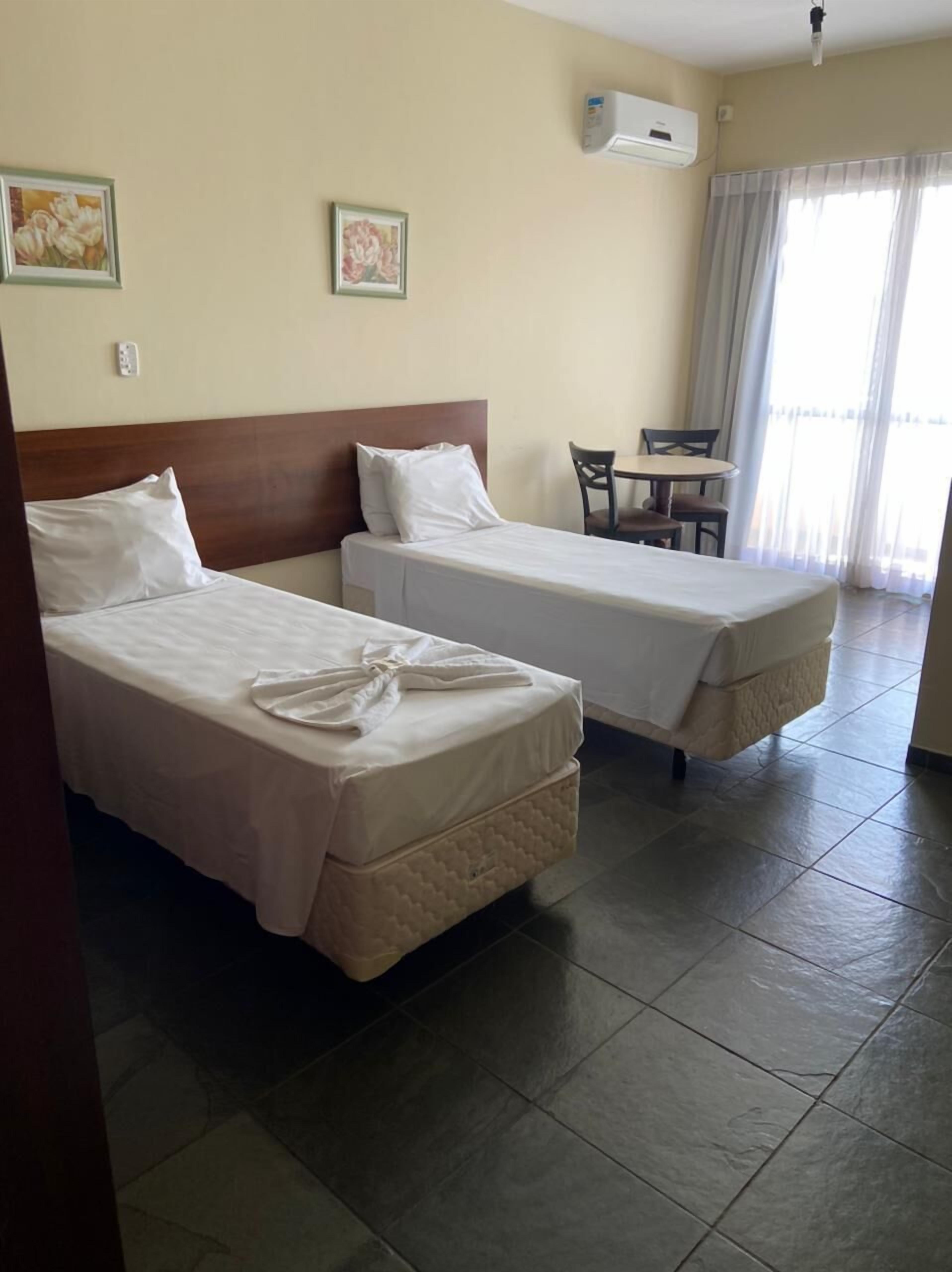 Habitación de hotel con dos camas individuales, una mesita con sillas, un equipo de aire acondicionado y una ventana con cortinas.