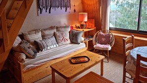 A cozy cabin interior with a wooden staircase, a sofa with patterned cushions, a small table with a tray, and a window with a view of trees.