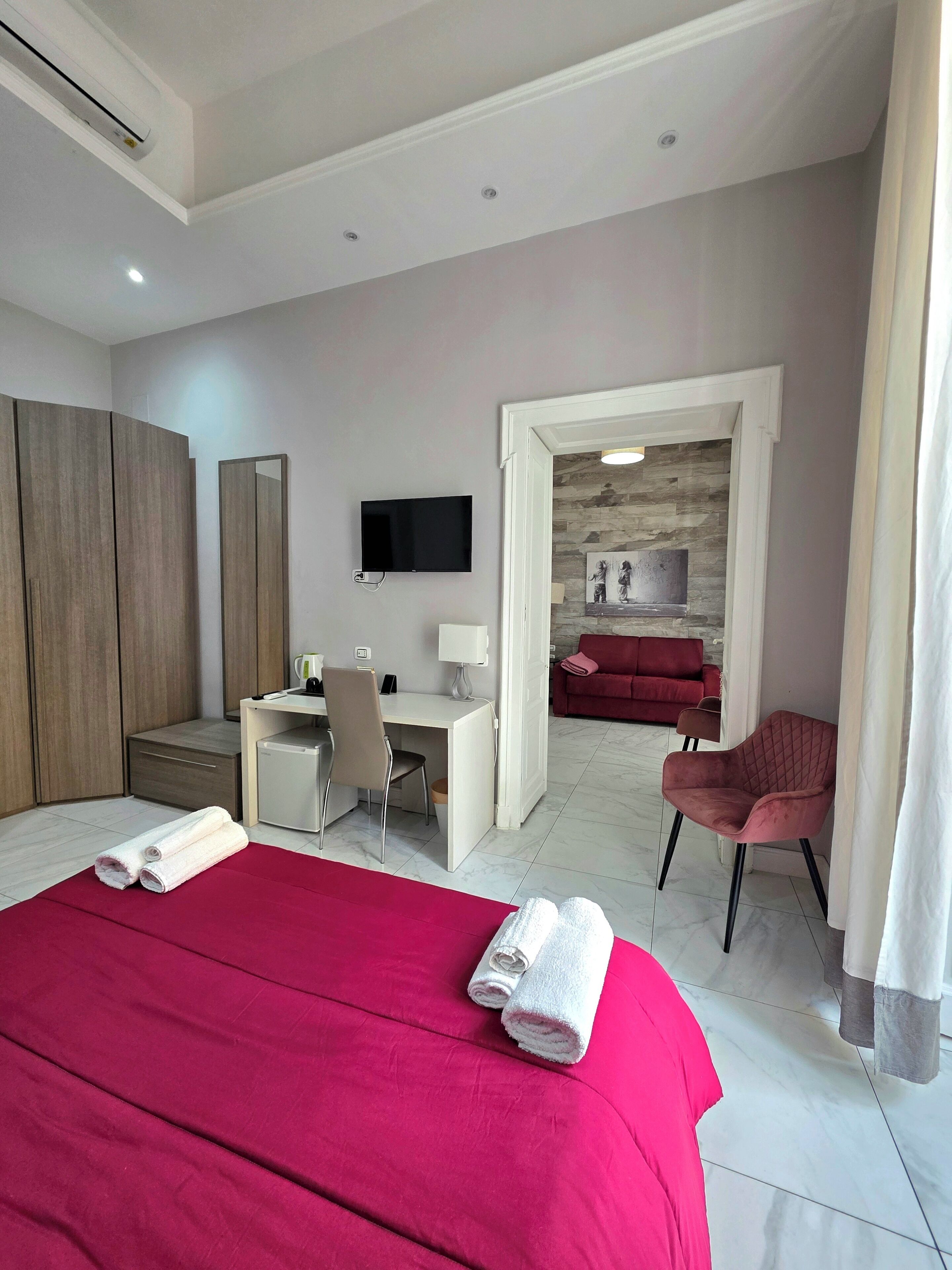 A hotel room with a bed, desk, chair, TV, and a view into a living area with a sofa and fireplace.