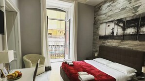 Une chambre d’hôtel avec un lit, un bureau et une chaise, une table à manger avec des fruits, et une vue sur le balcon extérieur.