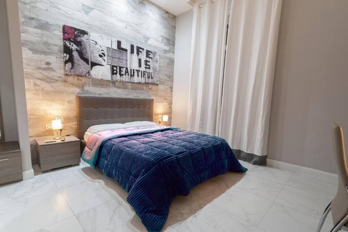 A bedroom with a bed, nightstands, a wall-mounted artwork, and a window with curtains.