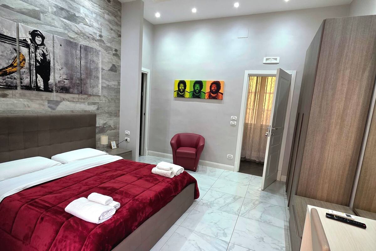 A modern hotel room with a large bed, a red blanket, a marble floor, a red armchair, and a wall with colorful artwork.