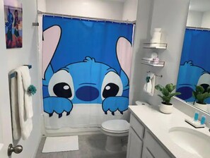 Ein Badezimmer mit Duschvorhang mit Stitch aus Lilo & Stitch, Toilette, Waschbecken mit Toilettenartikeln und Spiegel.
