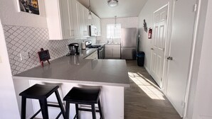 Moderni keittiö, jossa on valkoiset kaapit, keskellä oleva saareke jakkaroineen, jääkaappi ja pesuallas.