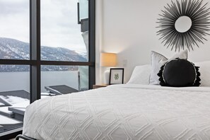 Una camera d'albergo con un'ampia finestra, un letto con copripiumino bianco, uno specchio a raggiera nero e un comodino con una lampada.