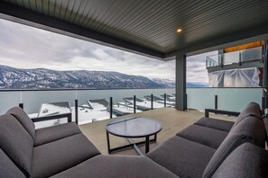A balcony with a view of snow-covered mountains, a glass table, and grey upholstered seating.