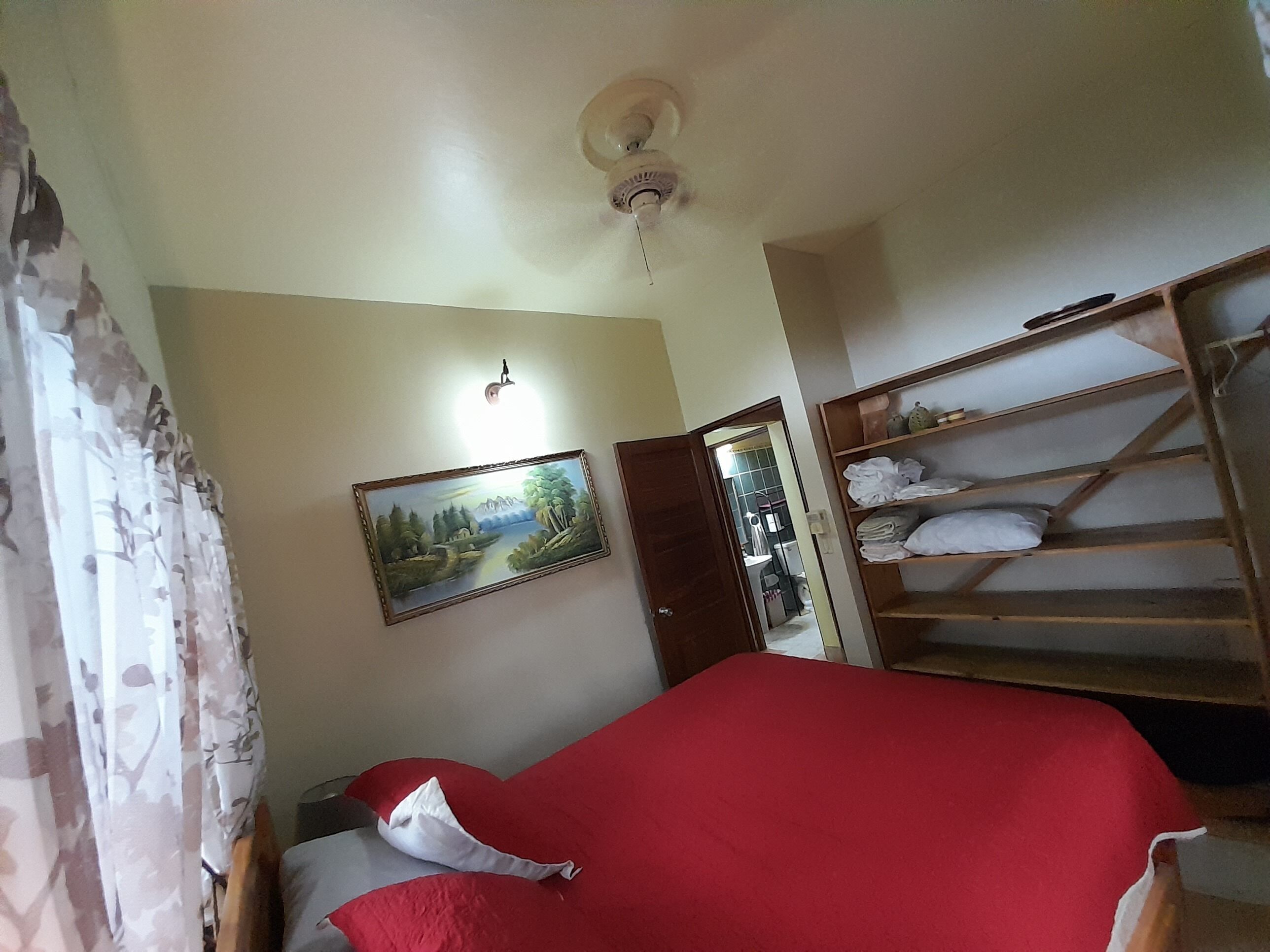 Un dormitorio con cama, un estante de madera con ropa doblada, un cuadro en la pared y un ventilador de techo.