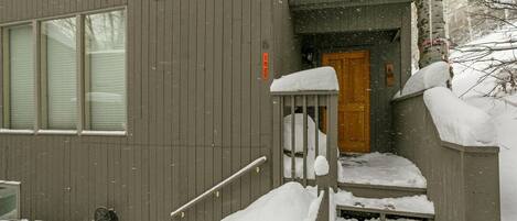 A wooden house with a wooden door and steps leading to the entrance. Snow is piled on the steps and around the house.