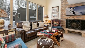 A cozy living room with a leather sofa, plaid cushions, a fireplace, and a large window overlooking a snowy landscape. - Wonderful Condo in the Aspens, Summer Pool & Tennis Courts (Vail)