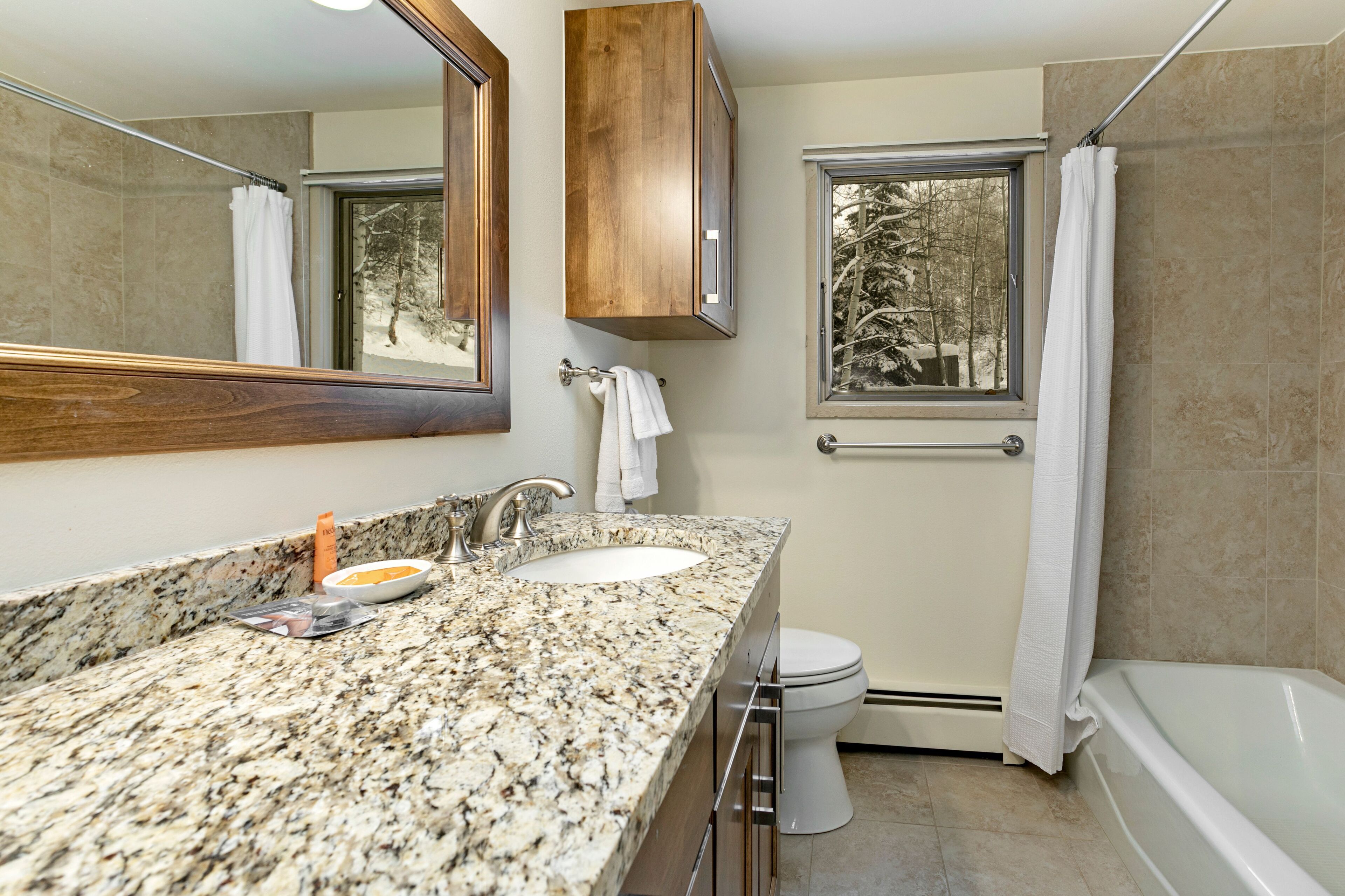 A bathroom with a granite countertop, two sinks, a toilet, a bathtub, and a window with a view of trees.