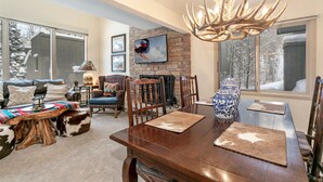 A cozy living room with a fireplace, a dining area with a wooden table, and a large window with a view of snow-covered trees. - Wonderful Condo in the Aspens, Summer Pool & Tennis Courts (Vail)